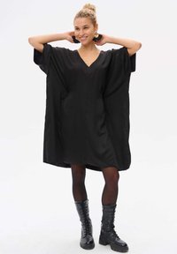 Schwarzes, übergroßes Kleid mit V-Ausschnitt, kurzen Ärmeln und lockerer Passform. Kombiniert mit schwarzen Schnürstiefeln im Combat-Stil. Glatte Stofftextur.