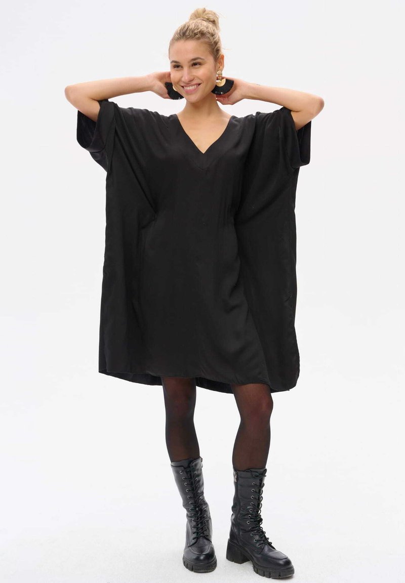 Schwarzes, übergroßes Kleid mit V-Ausschnitt, kurzen Ärmeln und lockerer Passform. Kombiniert mit schwarzen Schnürstiefeln im Combat-Stil. Glatte Stofftextur.