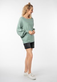 Hellgrüner, übergroßer Pullover aus weichem Material mit puffartigen Ärmeln, kombiniert mit schwarzen Denim-Shorts und weißen Sneakers.