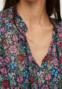 Bluse med blomsterdesign, der har en mørk baggrund med farverige rosa, blå og grønne blomster. V-hals-design i et blødt, let stof.