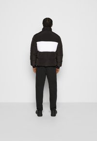 Hombre con el pelo trenzado, vistiendo pantalones negros, zapatos negros y una chaqueta acolchada negra con una amplia franja horizontal blanca, de pie de espaldas.