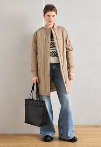 Μπεζ μακρύ μπουφάν bomber με ψηλό γιακά, σε συνδυασμό με πλεκτό με μοτίβο και καμπάνα τζιν σε μπλε χρώμα. Μαύρη τσάντα tote και σκουρόχρωμα παπούτσια ολοκληρώνουν την εμφάνιση.