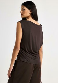 Brun ærmeløs top med draperet rygdetalje og løs pasform, lavet af blødt stof. Kombineret med mørkebrune bukser, minimalistisk design.
