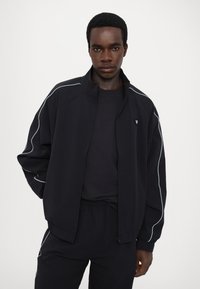 Svart zip-up jacka med vit pipingdetalj, texturerad tyg och avslappnad passform, kombinerad med en matchande svart skjorta och byxor.