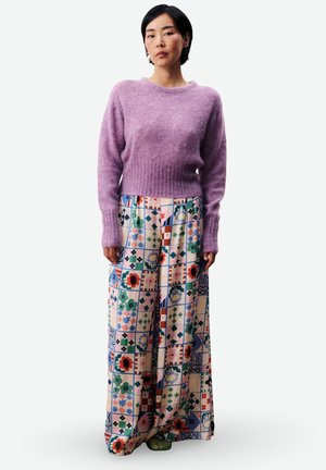 Personne portant un pull en laine lavande et un pantalon à jambe large avec un motif géométrique et floral multicolore, debout devant un fond blanc.