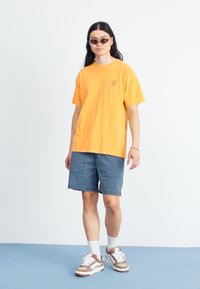Gul bomull t-skjorte, løs passform, korte ermer. Kombinert med blå shorts og brune joggesko. Tynne, ovale solbriller fullfører antrekket.