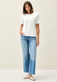 T-shirt bianco a maniche corte con un design grafico testurizzato sulla parte frontale, abbinato a jeans a gamba larga blu chiaro e sneakers beige.