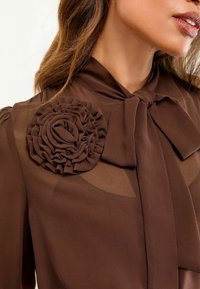 Braune Bluse mit einer Schleife und einem strukturierten Stoffblumendetail an der Schulter. Durchsichtige Abschnitte erhöhen die Eleganz des Designs.
