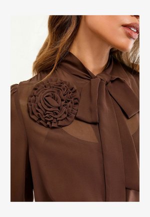 Pruun pluus, mis sisaldab nahaga lipsu ja tekstuuriga lilledetaili õlal. Läbipaistvad osad tõstavad disaini elegantsi.