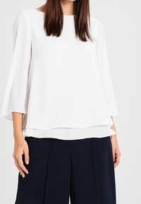 Weißes, fließendes Langarmblusenshirt mit rundem Halsauschnitt und geschichteter Saum. Hergestellt aus leichtem Stoff, passt gut zu navyblauen Hosen.