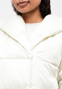 Λευκό μπουφάν με φουσκωτό σχέδιο, μεγάλο μυτερό γιακά και ομαλή γυαλιστερή υφή. Κλείσιμο με κουμπί και ελάχιστες λεπτομέρειες ραψίματος.