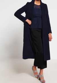 Lungo cappotto navy, top peplum navy texturizzato e pantaloni neri a gamba larga. Accentuato con tacchi grigi decorati da elementi circolari.