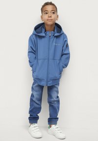 Blaue Kapuzenjacke mit Reißverschluss, strukturiertem Stoff, elastischen Bündchen und Seitentaschen; kombiniert mit blauen Jeans und weißen Turnschuhen.