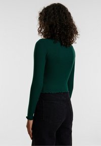Green ribbed long-sleeve top with a cropped design and scalloped hem, paired with black high-waisted jeans featuring back pockets.