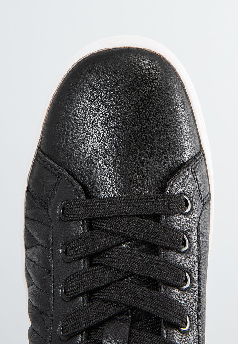 Vue en gros plan d'un bout de sneaker en cuir noir avec des lacets noirs et une semelle en caoutchouc blanche sur un fond clair.