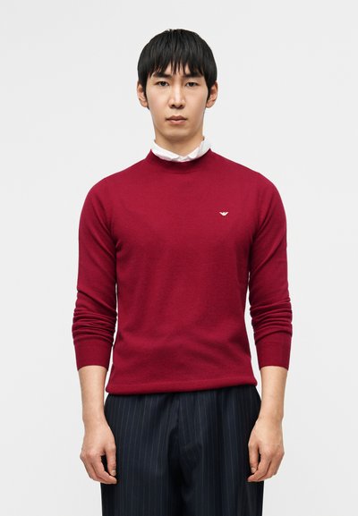 Pull en maille bordeaux avec col rond, manches longues et poignets côtelés. Petit logo sur la poitrine. Associé à un pantalon à rayures fines.