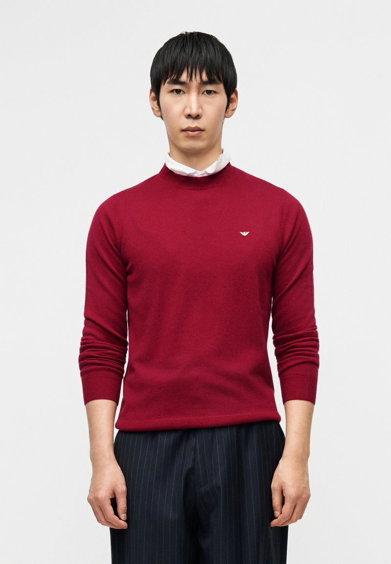 Μπορντό πλεκτό πουλόβερ με στρογγυλή λαιμόκοψη,  μακριά μανίκια και ριμπ μανσέτες. Μικρή λεπτομέρεια με λογότυπο στο στήθος. Συνδυάζεται με παντελόνια με λεπτές ρίγες.