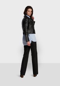 Black cropped leather jacket with zippers, worn over a light blue shirt, paired with black trousers and heels. Model poses from the side.