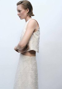Ensemble deux pièces beige pâle comprenant un top court sans manches et une jupe ajustée avec des détails texturés et des accents doux effilochés.
