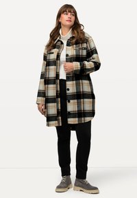 Schwarze und beige karierte Hemdjacke mit zwei vorderen Taschen, getragen über einem weißen Rollkragenpullover und kombiniert mit schwarzen Hosen und grauen Schuhen.