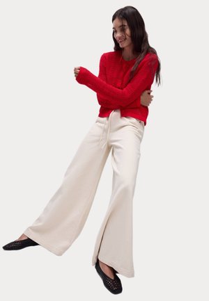 Cardigan rouge tricoté avec des manches longues et des boutons, associé à un pantalon large crème et des chaussures noires à bout pointu. Textures lisses.