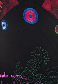 Černá látka s vyšitými vícebarevnými koncentrovanými kruhy a zeleným květinovým vzorem, obsahující částečný růžový text ve španělštině.