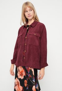 Bordeaux corduroy overhemdjack met een kraag, twee voorzakflappen en bruine knopen. De stof heeft een ribbelstructuur.