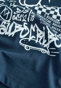 Tumši zila kokvilnas T-kreklu ar baltu grafisku tekstu un dizainiem, tostarp grafiti stilā rakstītiem burtiem un stilizētu skrituļdēļa ikonu.
