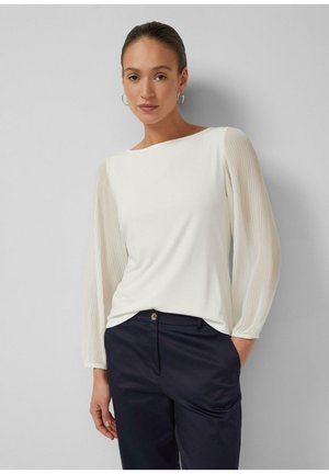 Vrouw draagt een witte blouse met doorzichtige geplooide lange mouwen en een zwarte broek, staat met één hand in de zak tegen een grijze muur.