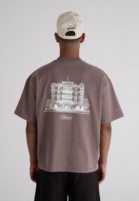 Brown oversized t-shirt made of cotton, featuring a white architectural print of a building and text. Back view, short sleeves.