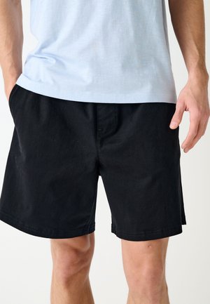 Mężczyzna ubrany w proste czarne szorty i jasnoniebieski t-shirt, z rękami przy kieszeniach, stojący na białym tle.