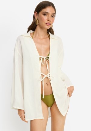 Woman wearing large sunburst earrings, olive green bikini, and a loose white shirt tied at front, standing against a plain light background.