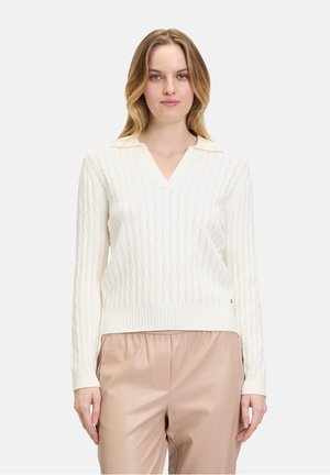 Cremfarbener Strickpullover mit V-Ausschnitt, ausgestattet mit einem gerippten Saum und Zopfmustern. Getragen mit beigen, locker sitzenden Hosen.