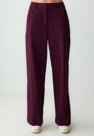 Pantaloni a gamba larga color bordeaux con una texture liscia, caratterizzati da una piega frontale e tasche laterali, abbinati a sneaker bianche.