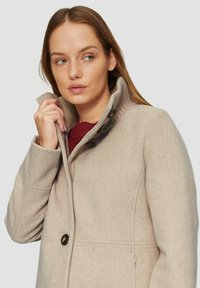 Beżowy wełniany płaszcz z żeberkowym kołnierzem i zapięciem na guziki, wykonany z teksturowanej tkaniny i posiadający boczne kieszenie. Bez wzorów ani ozdób.