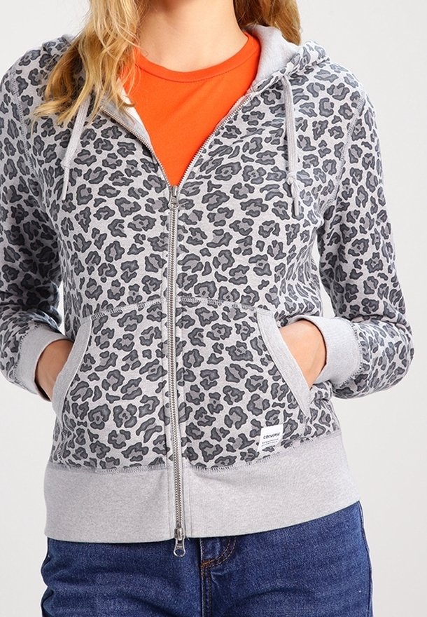 Femme portant un sweat à capuche gris zippé avec des taches léopard noires et un t-shirt orange en dessous, mains dans les poches, associé à un jean bleu.