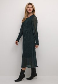 Pitkä, tummanvihreä mekko, jossa on smockattu yläosa, läpinäkyvät pitkät hihat ja röyhelökaulukset, yhdistettynä mustiin nilkkureihin.