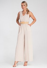 Beżowy kombinezon z dekoltem w kształcie litery V, szerokimi nogawkami i paskiem w talii. Wykonany z lekkiego materiału, z kieszeniami i zwiewnym fasonem.