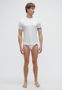 Hvid T-shirt med korte ærmer og sort elastikbånd med teksten "SQU" på armen, kombineret med hvide underbukser, båret af en stående mandlig model.