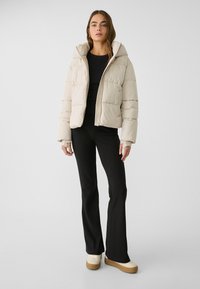 Beige pufferjacka med huva, med quiltade sektioner, dragkedja fram och ribbade muddars. Kombinerad med svarta tajta byxor och krämfärgade plattformsskor.