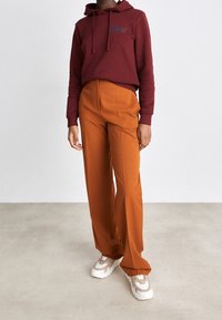 Sudadera burdeos con logo, pantalones naranja de pierna ancha y zapatillas beige. Tejido suave, corte relajado y detalle de capucha con cordón.