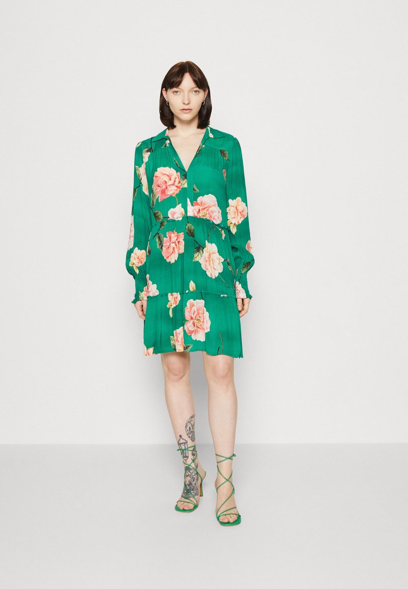 Grünes Blumenkleid mit langen Ärmeln, Falten und gestuftem Design. Es hat pinke und weiße Rosen und wird mit grünen Riemchenschuhen kombiniert.