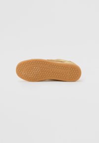 Gold sneaker with a textured surface, featuring a honeycomb-patterned rubber sole and a curved design. Bottom view highlights traction details.