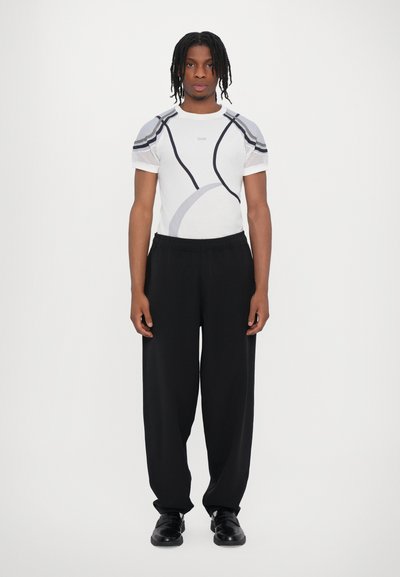 Weißes Sportshirt mit grauen Akzenten und Mesh-Ärmeln, kombiniert mit lockeren schwarzen Hosen und schwarzen Schuhen. Bequeme Passform und modernes Design.