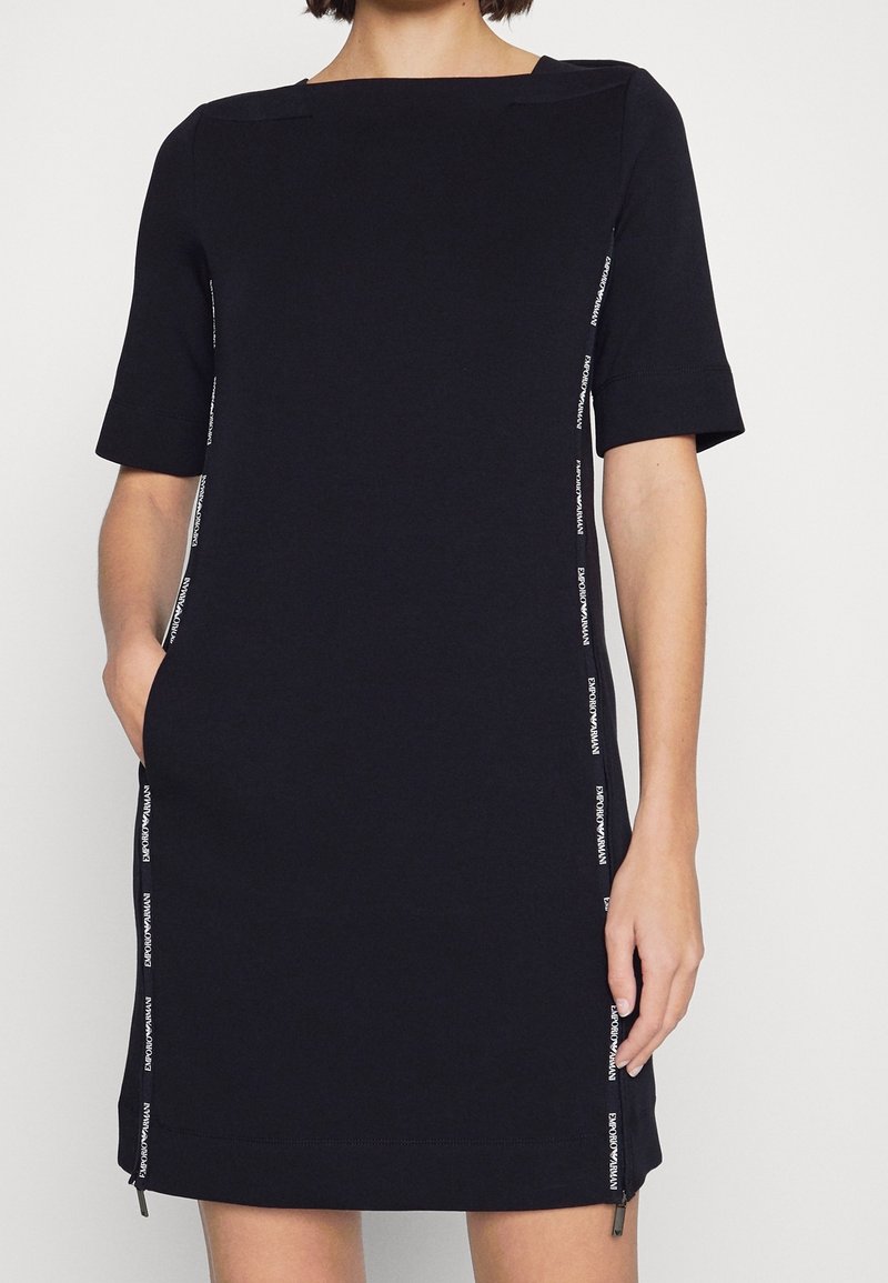 Černé šaty s krátkými rukávy, čtvercovým výstřihem, postranními kapsami a detaily loga podél stran. Vyrobeno z měkké, elastické tkaniny.