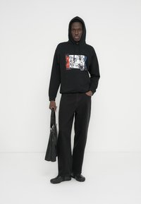 Melnā kapuce ar grafisko dizainu zilā, sarkanā un baltā krāsā; kopā ar melnām platu kāju biksēm un melnām kurpēm. Modele tur melnu somu.