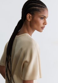 Femme aux longs cheveux tressés tirés en arrière, portant un haut beige à manches courtes, vue de profil sur un fond clair uni.