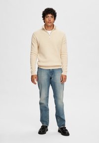 Hellbeige Strickpullover mit einem Viertelreißverschluss, gerippten Bündchen und Saum, kombiniert mit ausgewaschenen blauen Straight-Leg-Jeans und schwarzen Schuhen.
