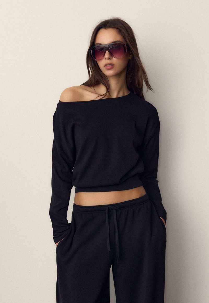 Femme portant des lunettes de soleil oversize, un haut noir à manches longues et épaules dénudées, ainsi qu'un pantalon noir avec cordon de serrage, posant les mains dans les poches sur un fond uni.