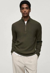 Pulover ribbed de culoare verde măsliniu cu guler înalt și închidere cu fermoar pe jumătate, suprapus peste o cămașă albă, asortat cu pantaloni de culoare crem.